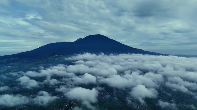Aerial view of Mount Gede Pangrango rising above a thick layer of clouds with lush green landscape and scattered settlements below in Sukabumi, West Java, Indonesia.