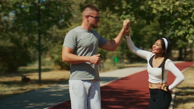 Two active individuals share a high-five while running on a track in a sunny park. They celebrate a moment of shared fitness and achievement outdoors.