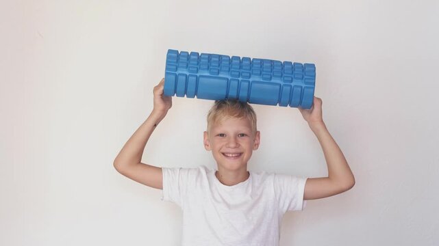 Victory and kids sport achievement, happy boy in white t-shirt holds blue foam roller over head with both hands on light background. Victory pose for sport stores and schools, boy smiles