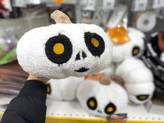Close-up of a plush white pumpkin toy with large black-and-yellow eyes, held in a hand at a store display. More Halloween-themed plush toys are softly blurred in the background. © Tatiana