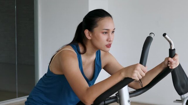 20s young adult southeast asian woman working out on stationary bike in indoor gym, dedicated and serious mood