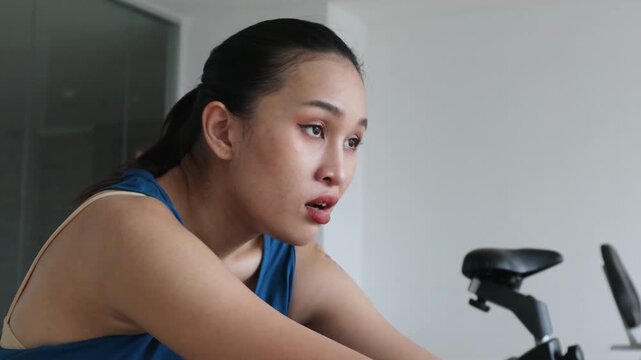 20s young adult southeast asian woman working out on stationary bike in indoor gym, panting, tired and exhausted, head and shoulder shot