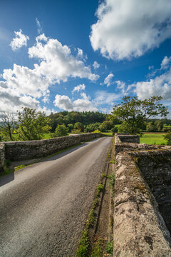 View of the historic 15th-century stone Horsebridge spanning the River Tamar, surrounded by lush green fields and trees under a bright blue sky in Horsebridge, England, United Kingdom.