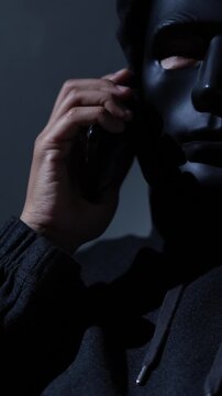Phone scammers protection. Unknown businessman wearing mask with covered face using mobile makes an anonymous call intimidating and threatening the interlocutor on dark background. hacker call center.