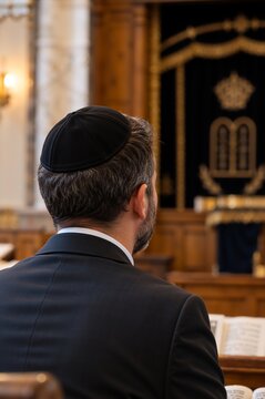 Adult Jewish man wearing black kippah head covering during religious service in synagogue. Rear view of head and shoulders. Focus on traditional attire and place of worship. Copy space