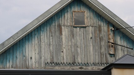 A close-up of the wooden gable of an old village house. The attic facade is clad in vertical gray boards with traces of turquoise paint. A small square window is located in the center © Skyliz