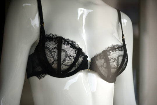 Close-up of a black underwire bra featuring sheer floral lace, thin shoulder straps and a gold central detail, displayed on a white mannequin torso in a shop window.
