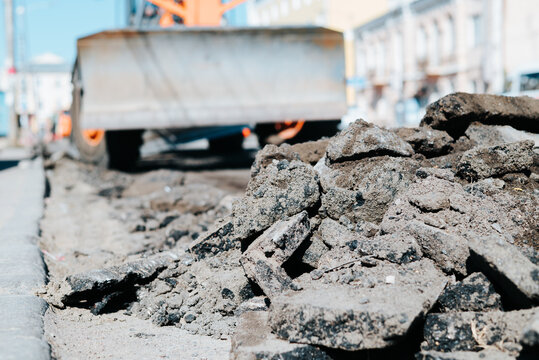 Pieces of broken asphalt are scattered on the ground at a construction site. Heavy machinery is visible in the background, indicating ongoing urban roadwork