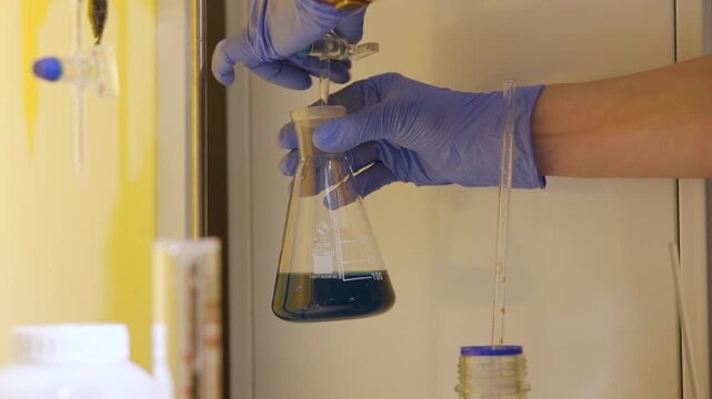 Gloved hands adding reagent drop into Erlenmeyer flask with blue solution inside laboratory fume hood, droplet poised at dropper tip