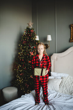 Kid girl 7-8 year old wearing red checkered pajamas holding Christmas present box over decorated eve in bedroom. Childhood. Winter holidays.