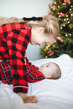 Child girl playing with baby in bed wearing red pajamas over Christmas tree at background. Winter holiday season. Childhood.