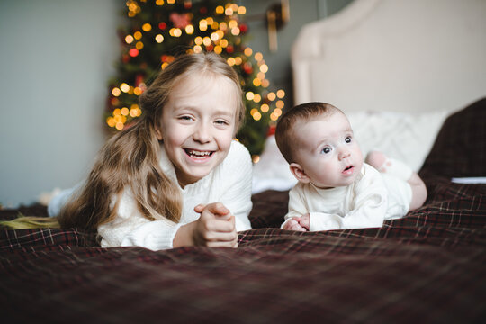 Two smiling children wearing knitted sweaters having fun in bed over Christmas tree at background in room. Winter holiday season.