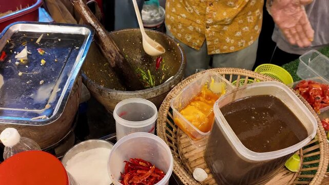 Close up shot of street food vendor hands in gloves adding palm sugar paste and seasonings into a clay mortar for traditional Thai papaya salad.
