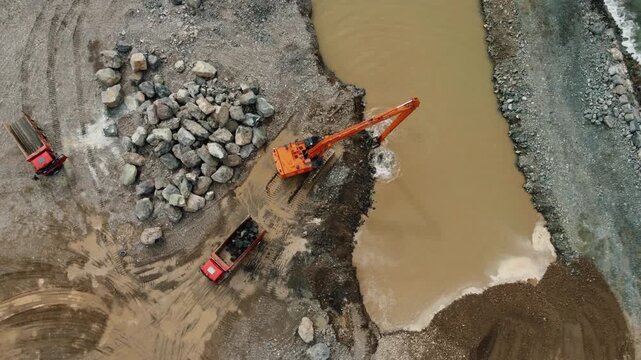 Aerial top-down view of a construction site where an excavator is dredging mud from a river, loading dump trucks while a wheel loader moves earth on the bank in an industrial quarry