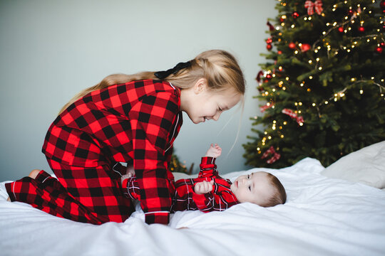Two children playing in bed wearing red checkered pajamas having fun over Christmas tree at background in room. Winter holiday season.