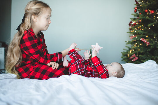 Child girl playing with baby in bed wearing red pajamas over Christmas tree at background. Winter holiday season. Childhood.
