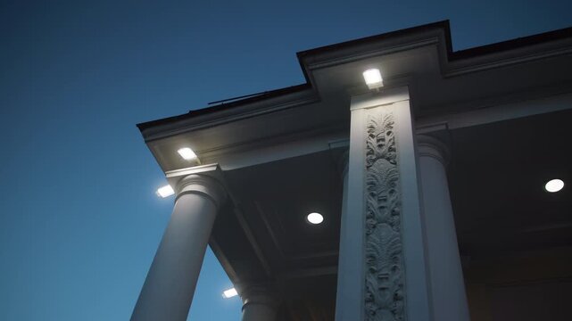 Low angle dusk view of illuminated classical portico with paired columns, carved pilaster details, and recessed ceiling lights under ornate entablature and cornice