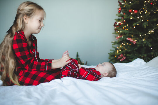 Two children playing in bed wearing red checkered pajamas having fun over Christmas tree at background in room. Winter holiday season.