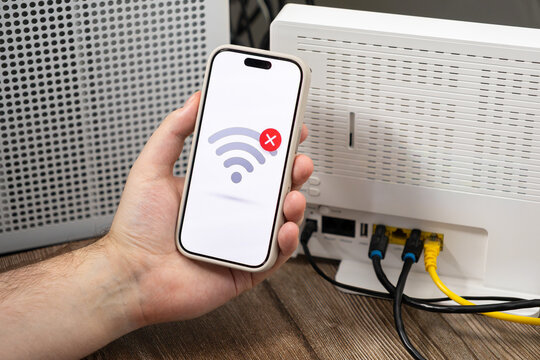 Smartphone held by a person displays a no signal icon near a wireless router, capturing technical difficulties with network connectivity issues in a modern home office environment setup