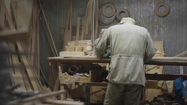 Woodworker leaning over cluttered workbench in dim carpentry workshop, focused on stacked cut timber beside power tool, corrugated wall with peeling paint and shelves