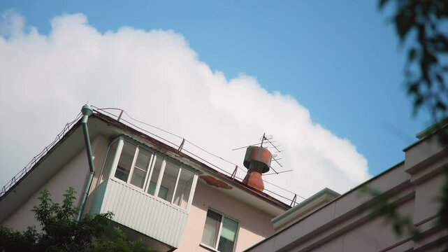 Low-angle view of pastel residential rooftop with balcony, chimney cap, rooftop antenna, and metal safety railings against blue cloudy sky