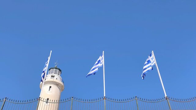 View of Greek flags waving in the breeze, with the historic Lighthouse of Alexandroupoli standing prominently in the background against a brilliant blue sky in Alexandroupoli, Thrace, Greece.