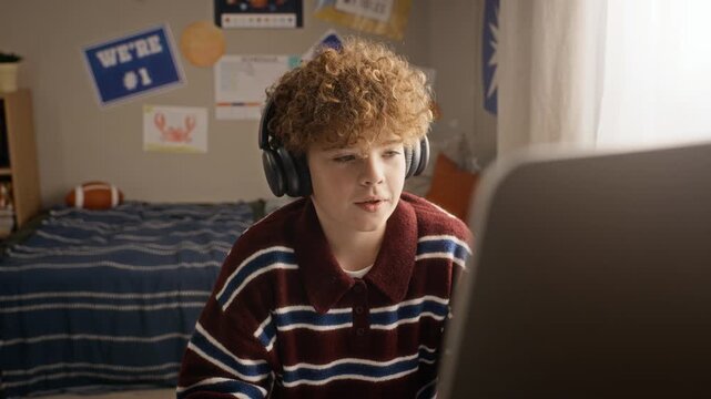 Caucasian teenager wearing headphones writing notes during online lesson on computer with camera zooming out. Footage suitable for e-learning platforms, distance education, edtech advertising