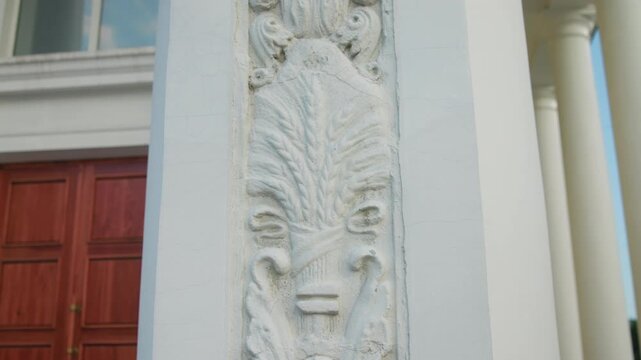 Ornate carved stone pilaster with leaf and scroll relief at exterior building entrance beside wooden doors and painted columns under soft daylight in warm tones