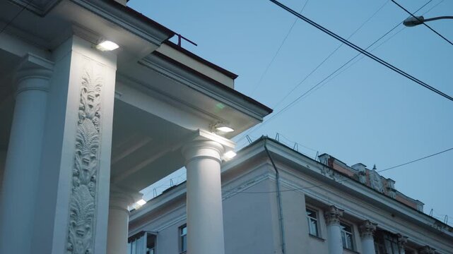 Low angle dusk view of classical building entrance with illuminated white columns, ornate pilaster relief, projecting canopy lights, and overhead utility wires
