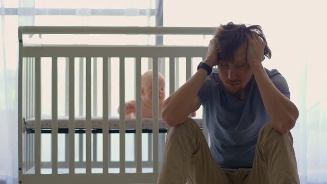 Sad and exhausted father sitting beside crib with five-month-old son inside. Concept of parental exhaustion.