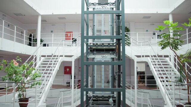 Framed view of glass elevator shaft in bright modern office atrium with symmetrical white metal staircases, open walkways, potted plants, and glossy tile flooring