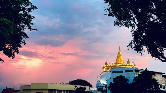 Golden Mountain Pagoda at Wat Saket standing beneath a vibrant pastel sunset sky with pink and blue clouds in Bangkok, Thailand, framed by silhouettes of lush green tree leaves in the evening.