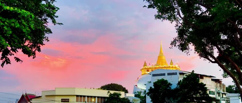 Golden Mountain Pagoda at Wat Saket standing beneath a vibrant pastel sunset sky with pink and blue clouds in Bangkok, Thailand, framed by silhouettes of lush green tree leaves in the evening.