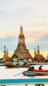 Wat Arun pagoda against a dramatic orange sunset sky over the Chao Phraya River in Bangkok, Thailand, creating a serene, golden twilight scene along the historic waterfront.