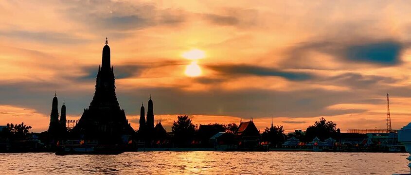 Silhouette of Wat Arun pagoda against a dramatic orange sunset sky over the Chao Phraya River in Bangkok, Thailand, creating a serene, golden twilight scene along the historic waterfront.