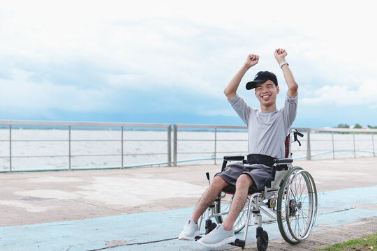 Excited disabled Asian teen in wheelchair wearing cap aising arms in celebration at lakeside park, inclusive outdoor lifestyle.