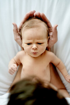 Baby, above and hands of parent in nursery with bonding, frown or serious face with head support. Family house. people and early child development with connection, scowl or grumpy infant in home
