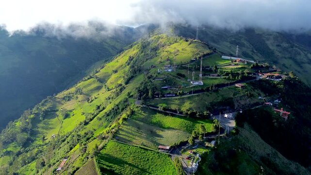 Drone footage presents a broad aerial view of a dark volcanic peak above a belt of clouds, with green slopes and patchwork fields across the foreground. Bright clouds frame the mountain beneath a deep