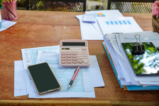Tax accounting and financial planning with calculator, 1040 form, and smartphone on wooden desk. Report, paperwork, pencil, and binder clip in business workspace.