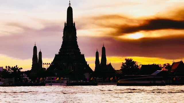 Silhouette of Wat Arun pagoda against a dramatic orange sunset sky over the Chao Phraya River in Bangkok, Thailand, creating a serene, golden twilight scene along the historic waterfront.