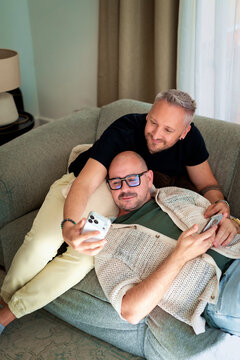 Same-sex male couple chilling on the couch in their living room, looking at their smartphones