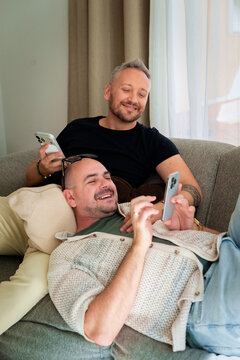 Same-sex male couple chilling on the couch in their living room, looking at their smartphones