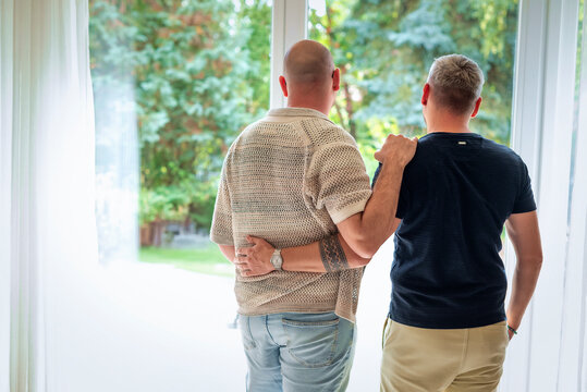 Rear view of a same-sex male couple standing by a floor-to-ceiling window, looking out and daydreaming.