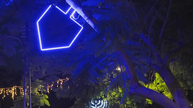 Blue neon dreidel and a white menorah light installation hanging from a tree for the jewish holiday of Hanukkah, celebrating the festival of lights with urban decorations in a park
