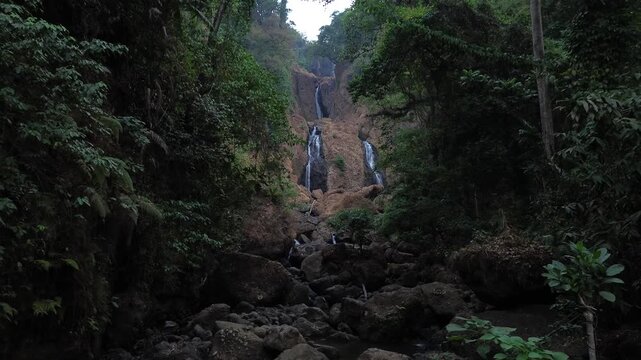 Aerial drone footage of a scenic waterfall flowing over rocky terrain through a dense tropical forest. Multiple cascading streams descend between mossy rocks and lush green vegetation.