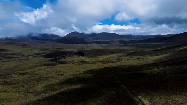 Drone footage looks down over cattle grazing in patchy green pasture on a rural hillside, with a winding road, utility poles and a small settlement in the surrounding mountain valley. Filmed in Tungur