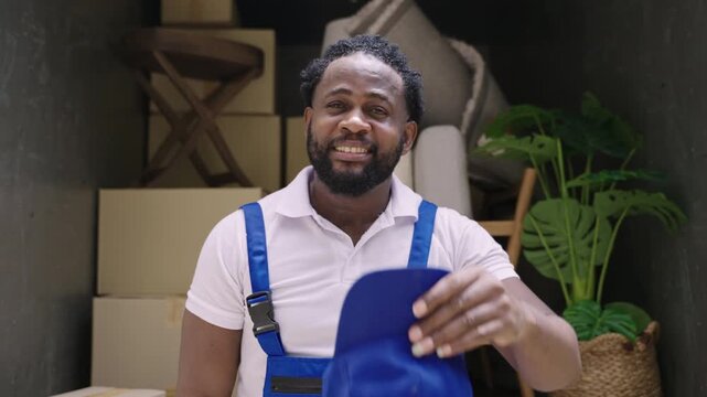 Happy professional African American man, mover and delivery service, in a company uniform, standing, smiling, and taking off a blue cap while looking at the camera, relaxing after loading onto truck. 