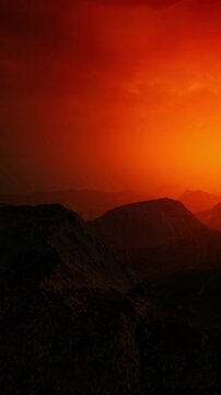 A sunset paints the sky orange as it sets behind distant mountains. The landscape is mostly dark with rocky features and a hazy atmosphere. The scene captures the end of the day.