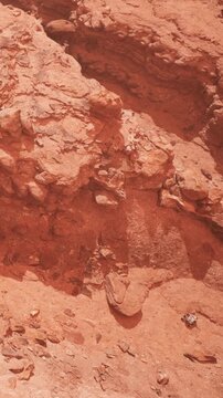A rocky surface shows various shades of red with rough textures. Small stones are scattered among the larger rock formations. This scene is taken in a desert area under clear blue sky.