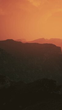 The sun sets behind the mountains, creating an orange sky. Silhouetted peaks are visible against the colorful backdrop. The view captures the beauty of nature at dusk.